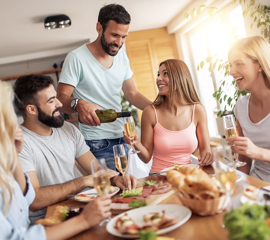group of friends at kitchen table. A man pours wine for a girl in a pink tank top.